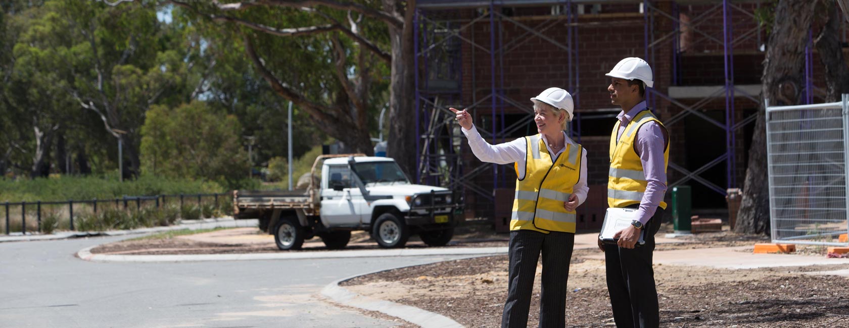 Two people in safety vests and helmets discussing plans at a worksite, with a utility vehicle and building structure in the background.
