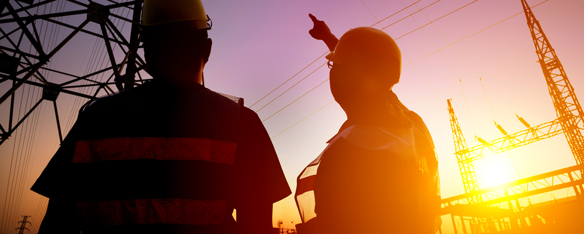 Two workers in safety gear inspecting powerlines at sunset, with transmission towers and electrical infrastructure in the background.