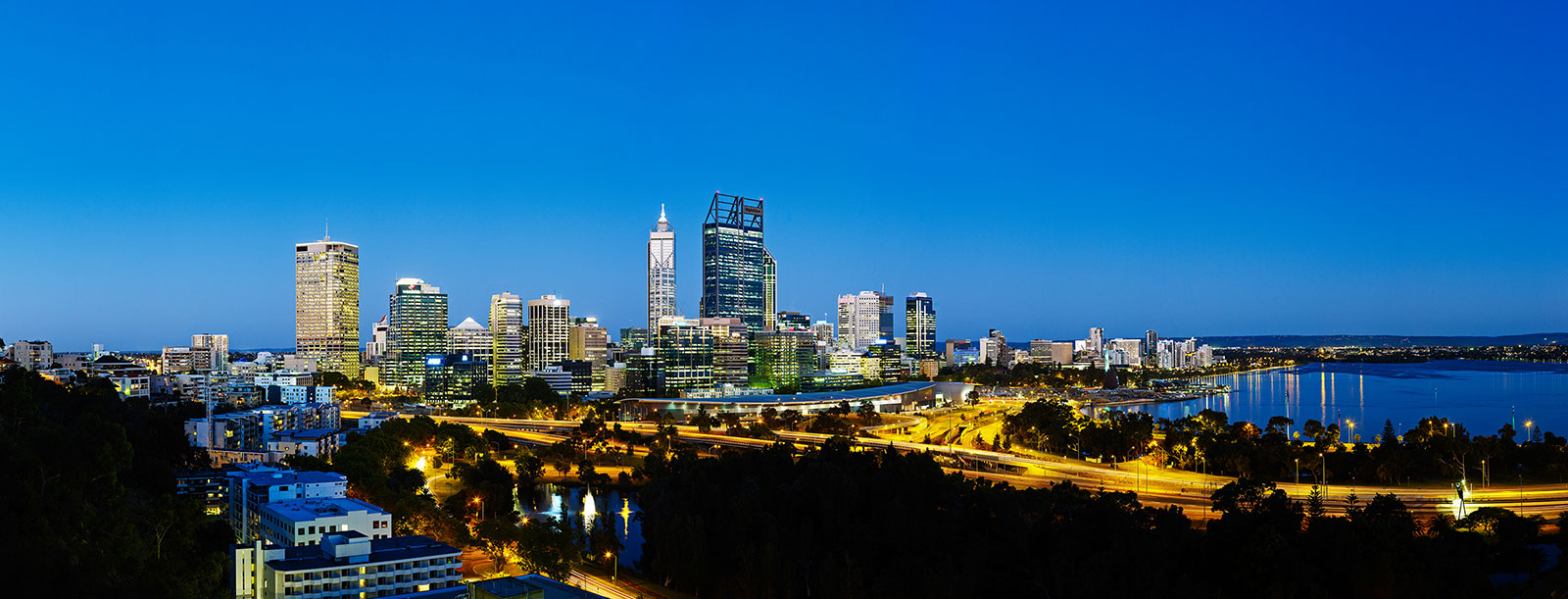 Panoramic view of the Perth city skyline at dusk, with illuminated buildings and roads reflecting on the Swan River