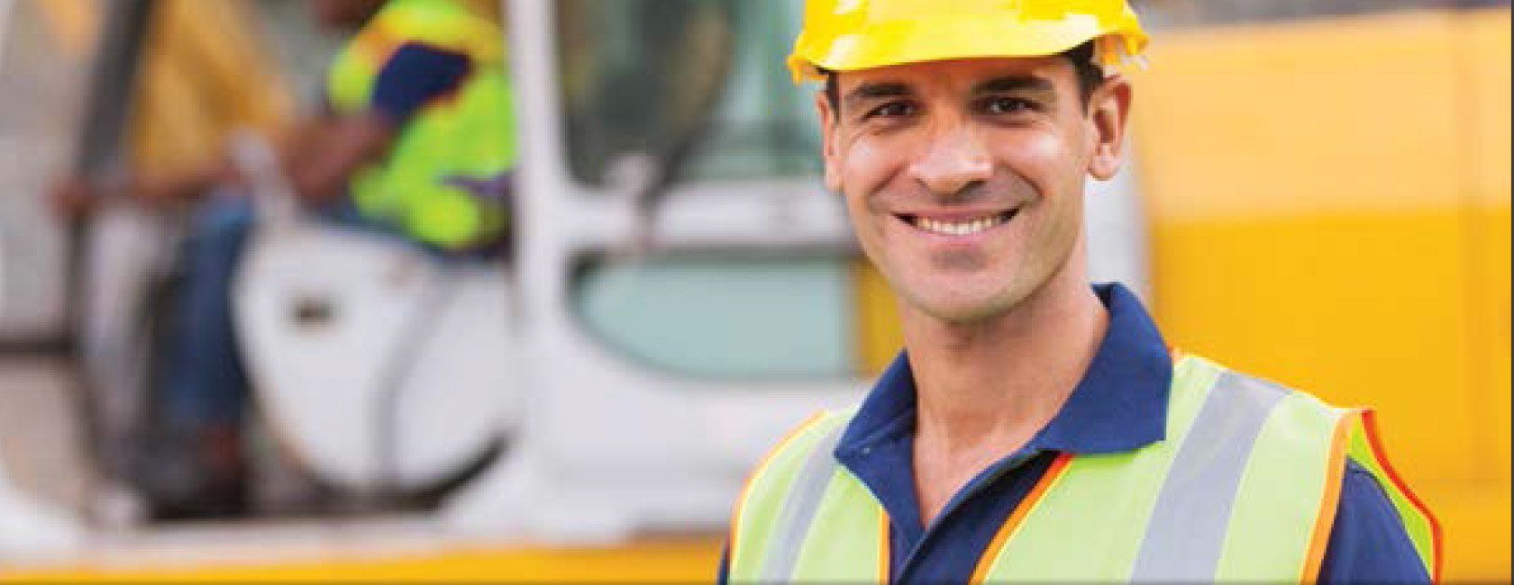 A worker wearing a yellow hard hat and reflective safety vest, standing in front of construction equipment with another worker in the background.