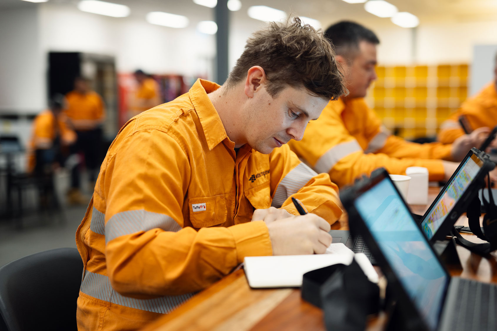 A Western Power apprentice in an orange high-vis shirt writes in a notebook at a table with laptops and colleagues nearby.