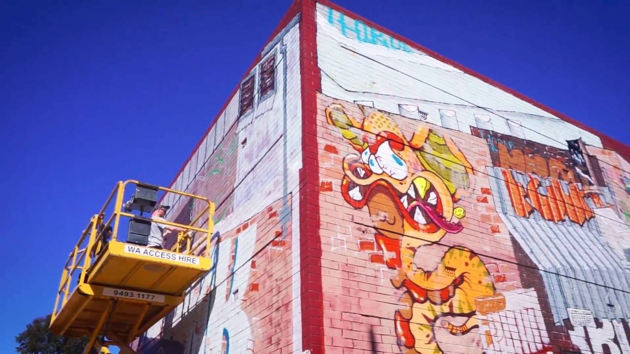 An artist on an elevated work platform next to a commercial building with urban artwork.