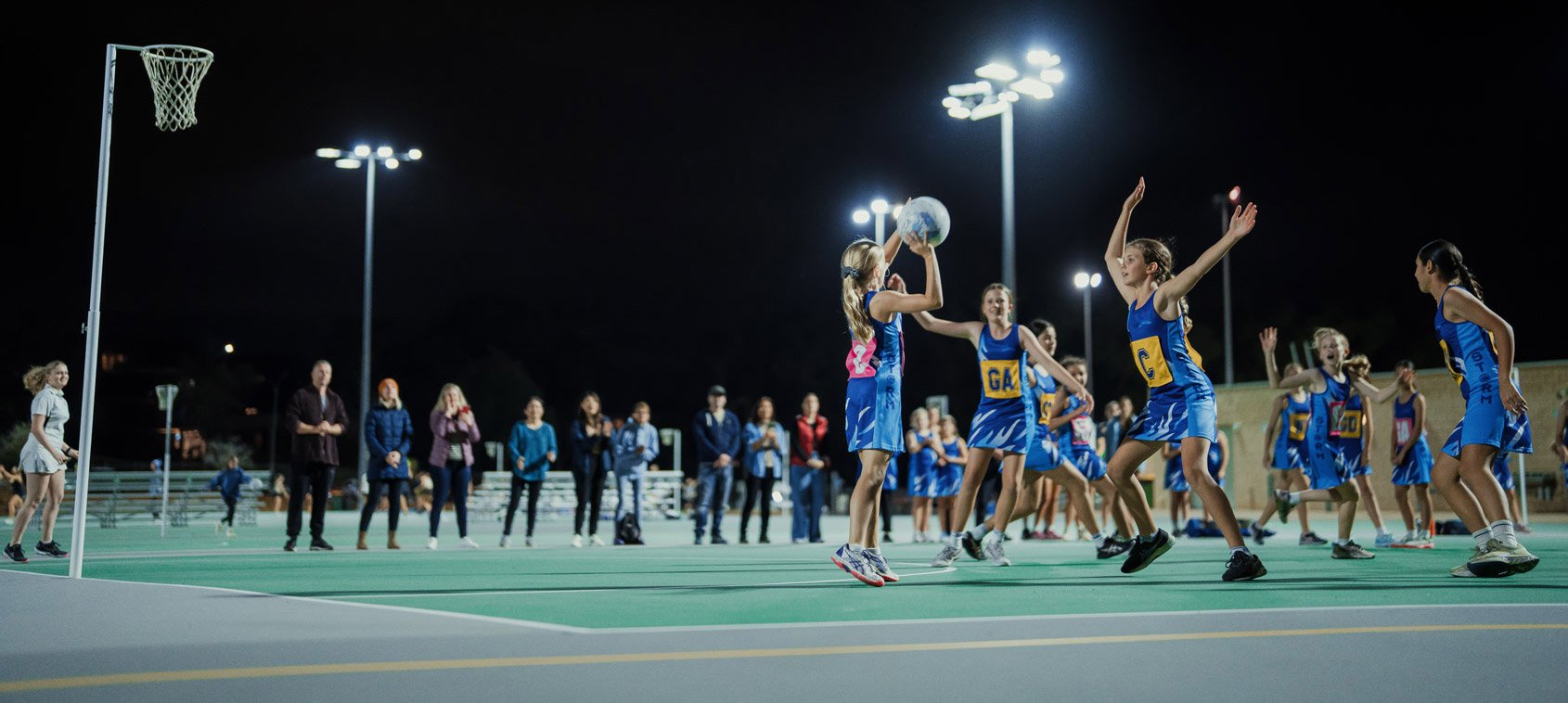 A netball game in progress at night being lit by floodlights with parents watching on the sidelines